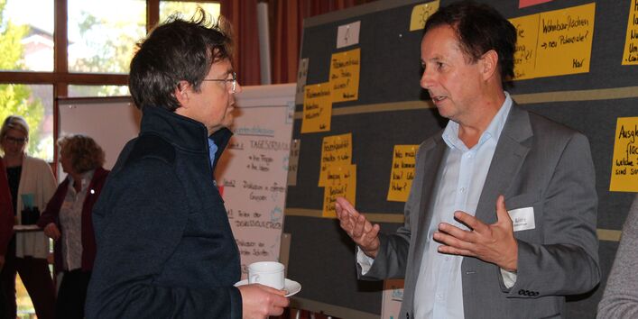 Zwei Teilnehmende unterhalten sich gestikulierend miteinander. Sie stehen vor einer Moderationswand, ein Flipcharts und andere Teilnehmende sind im Hintergrund sichtbar. Der Blick geht links durch das Fenster hinaus ins Grüne.