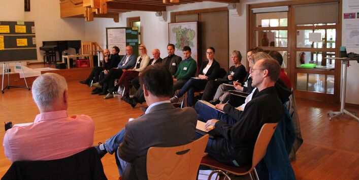 Teilnehmende sitzen im Stuhlkreis mit übergeschlagenen Beinen. Sie halten Notizpapier und Stifte in den Händen. Im Raum verteilt stehen Flipcharts und Moderationswände mit Kartenmaterialien darauf. Über den Köpfen der Teilnehmenden befindet sich die hölzerne Treppengalerie und darunter ein verlaster Durchgang.