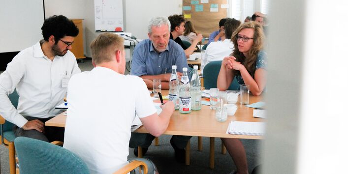 Planspiel: Teilnehmende sitzen an Gruppenarbeitstischen und denken nach. Im Vordergrund schreibt ein Teilnehmer im weissen T-Shirt etwas auf eine hellblaue Moderationskarte.