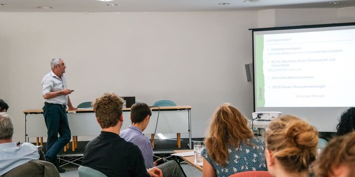 Vortragender zeigt seine Präsentation im lockeren Stil. Er steht mit einer Hand auf einen Tisch gestützt und mit der anderen Hand hält er einen Pointer. Mit dem Pointer zeigt er seine Präsentation auf einer Leinwand im Hintergrund. Im Vordergrund sitzen die Teilnehmenden und hören.