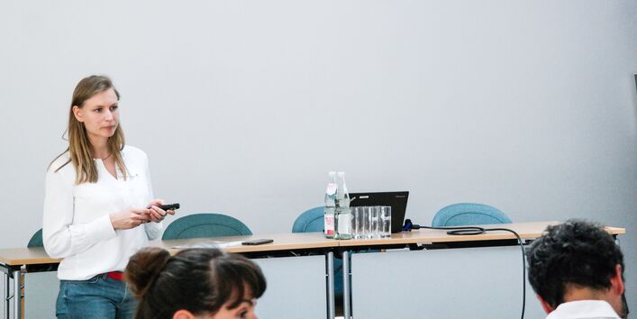 Vortragende steht vor einem Podiumtisch und hält einen Pointer in ihren Händen. Auf dem Podiumstisch stehen Gläser und Getränkeflaschen.