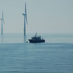 Das Foto zeigt ein Schiff vor drei Windenergieanlagen mit einem Monopile-Fundament. Davor sieht man ein im Verhältnis sehr klein wirkendes Schiff.