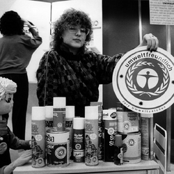 Eine Frau hält das Umweltzeichen in die Kamera, vor ihr auf einem Tisch viele Produkte, die das Umweltzeichen tragen, zum Beispiel Rasierschaum-Flaschen.