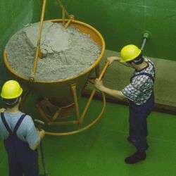Zwei Männer mit Arbeitskleidung und Sturzhelmen stehen in einem tiefen grünen Teichbehälter mit einem von oben herabhängenden, trichterförmigen Gefäß mit Sand
