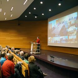 Barbara Hendricks hält im Hörsaal des UBA eine Rede, im Hintergrund sieht man auf einer Leinwand die Videoübertragung an andere Standorte. Im Vordergrund das Publikum in Dessau.
