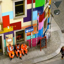 Drei Männer in den orangenen Anzügen der Müllabfuhr sitzen auf einer Bank vor einem bunt gestrichenen Eckhaus und trinken Kaffee