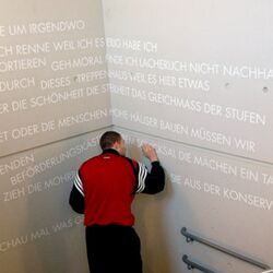 Ein Mann schreibt mittels besagter Schablonen weitere Begriffe und Worte mit weißer Farbe an die Betonwand