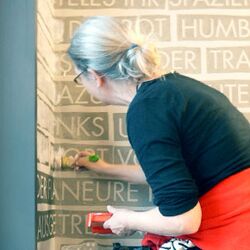 Christiane Keppler streicht mittels weißer Farbe durch die Wortschablonen ihre Texte an die Wand des UBAs Treppenhaus