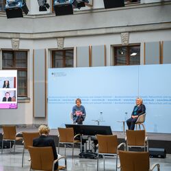 In dem hellen historischen Innenhof sitzen 2 Frauen auf Schalenstühlen auf dem Podium vor einem blauem Hintergrund. Links davon steht ein großer flacher Bildschirm, der die zugeschalteten Konferenzteilnehmenden zeigt. Vor dem Podium stehen leere Stühle. Auf einem Stuhl beugt sich eine Teilnehmerin über Ihre Unterlagen.