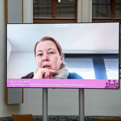 In dem hellen historischen Innenhof zeigt sich eine Teilnehmende auf dem großen flachen Bildschirm sprechend und nachdenklich. Sie hält sich die rechte Hand mit leicht gebeugten Fingern vor das Kinn.