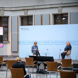 Der Blick geht in dem hellen historischen Innenhof von hinten über das Publikum hin zum Podium vor blauem Hintergrund, wo die Umweltministerin und eine Konferenzteilnehmerin sitzen im Gespräch mit dem Präsidenten des Umweltbundesamtes, der links des Podiums auf dem großen flachen Bildschirm zugeschaltet wurde.