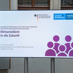 Der Blick geht auf das Konferenzlogo, welches sich im hellen historischen Innenhof auf dem großen flachen Bildschirm nahe zeigt