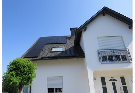 Ein weißes Einfamilienhaus mit großem Erker und PV auf dem schwarzen Dach vor blauem Himmel..