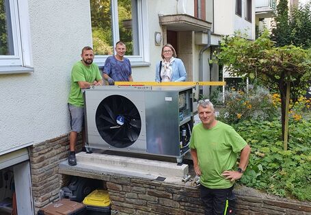 Eine Wärmepumpe wird rechtwinklig zu einem Mehrfamilienhaus auf einer Mauer zur Kellereinfahrt montiert, daneben stehen Mitarbeiter der Installationsfirm und die Bürgermeisterin