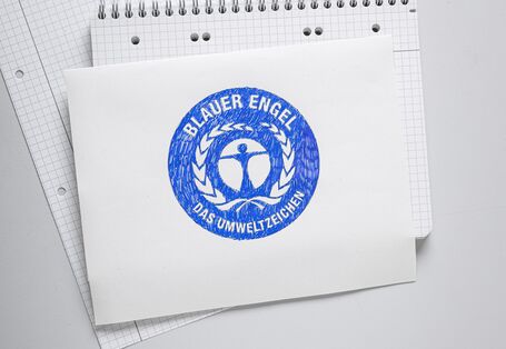 Foto von oben. Auf einem Schreib-Block liegt ein weißes Blatt Papier. Darauf ist das runde, blaue Logo vom „Blauen Engel“ mit blauem Stift gemalt. Ganz oben liegt ein zusammengeknülltes Stück Papier.