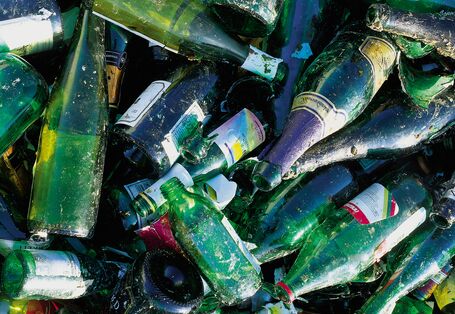 A high-angle, close-up shot of a pile of discarded green and dark glass bottles intended for recycling. The bottles, mostly wine and champagne varieties, are cluttered together, some broken, with visible paper labels and residue, showing textures of dirt and wear.