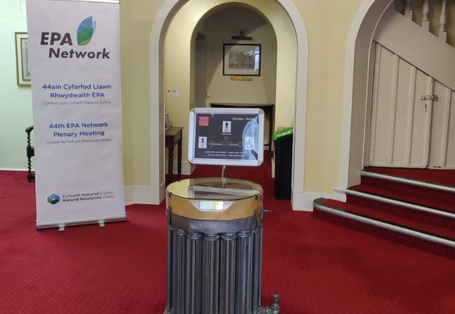 Entrance hall with a red carpet. In the foreground a monitor displaying info and on the left a rectangular poster announcing a meeting.