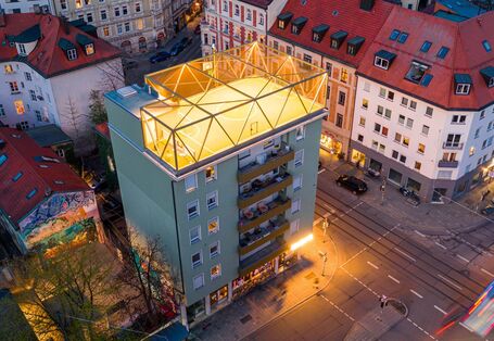 Luftbild eines Gebäudes an einer urbanen Straßenkreuzung in einer Abendstimmung. In der Bildmitte hebt sich ein beleuchteter Sportplatz auf dem Gebäude ab.