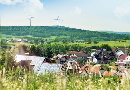 Ein Foto mit Wiese und Einfamilienhäusern im Vordergrund. Auf manchen Dächern befinden sich Solaranlagen. Im Hintergrund befindet sich landwirtschaftliche Fläche, bewaldete Hügel und drei Windkraftanlagen. 