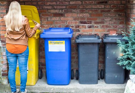 A person with long blonde hair, wearing a brown leather jacket and blue jeans, stands with their back to the camera while disposing of a metallic object into a row of recycling bins. The bins are lined up against a red brick wall. From left to right, there is a large yellow bin with its lid open, a blue bin labeled "Altpapier" (waste paper), and two smaller dark grey bins. The scene is outdoors on a paved surface.
