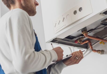 Foto. Ein Handwerker in blauer Arbeits-Hose steht vor einer weißen Gas-Therme an der Wand. Er schaut auf die Rohre unter dem Gerät und schreibt etwas auf ein Klemm-Brett.