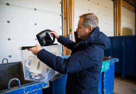 Foto. Ein Mann in einer blauen Winter-Jacke steht vor einem großen, blauen Müll-Container. Er hält eine durchsichtige Kiste aus Plastik. Darin liegt ein weißer Wasser-Kocher. Er legt ein schwarzes Elektro-Gerät in den Container.