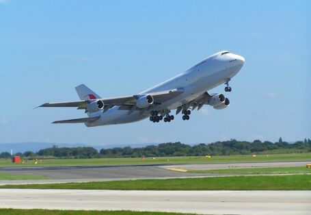 A large white Boeing 747 jumbo jet taking off from an airport runway against a clear blue sky, with its landing gear still extended.