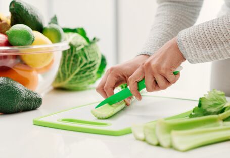 Foto. Ein Mensch schneidet grünen Sellerie auf einem Schneide-Brett. Daneben steht eine große Schüssel mit buntem Obst und Gemüse, wie Avocados, Zitronen und Äpfeln.