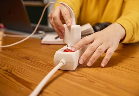 A person’s hands are shown plugging a white power adapter into a white power strip on a wooden table. The power strip has a red illuminated power switch.