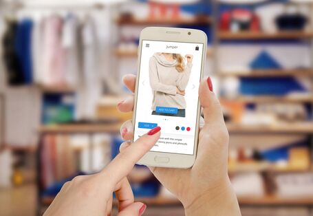 Foto. Zwei Hände halten ein weißes Smart-Phone. Auf dem Bild-Schirm ist ein beige-farbener Pullover in einem Online-Shop zu sehen. Ein Finger mit rotem Nagel-Lack tippt auf den Bild-Schirm. Der Hintergrund ist unscharf. Man erkennt Kleider-Ständer in einem Geschäft.