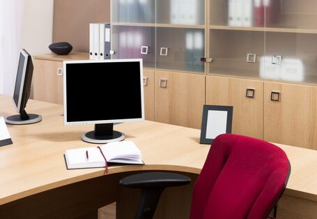 Ds Bild zeigt einen Büroarbeitsplatz mit Schränken im Hintergrund und einem Monitor auf dem Schreibtisch.