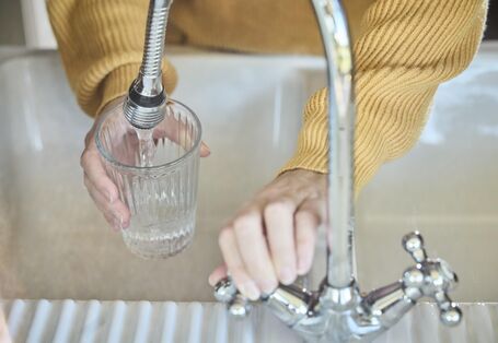 High-angle, close-up shot of hands wearing a yellow sweater, holding a clear ridged glass and filling it with fresh water from a modern chrome kitchen faucet.
