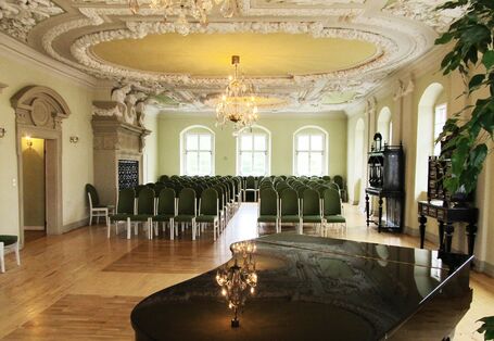 Barocksaal mit Stuckdecke und Holzparkettboden, im Vordergrund steht ein schwarzer Flügel, der Raum ist bestuhlt