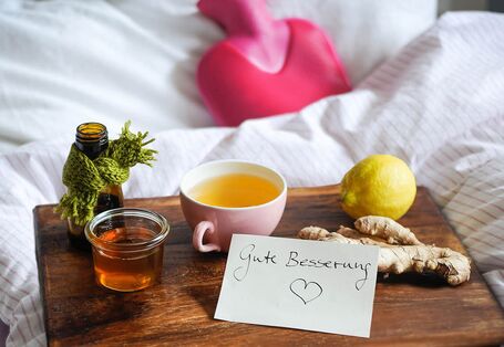 Foto Bett mit Wärmflasche und Tablett, darauf Honig, Tee, Ingwer, Zitrone und Schild mit „Gute Besserung“