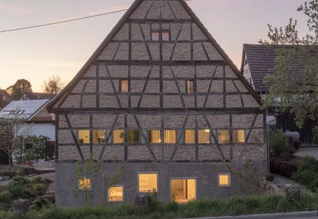 : Foto der Giebelwand eines viergeschossigen Fachwerkhauses mit Gefachen aus Feldstein und Satteldach. In der dörflichen Szene während der Dämmerung sind die Fenster mit warmem Licht erleuchtet.