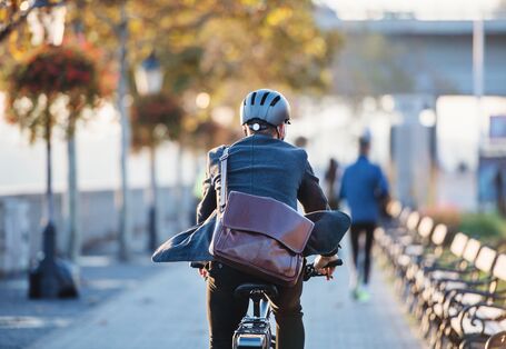 Ein Mann in einem Anzug und mit Aktentasche, von hinten fotografiert, fährt mit dem Fahrrad eine Straße entlang.