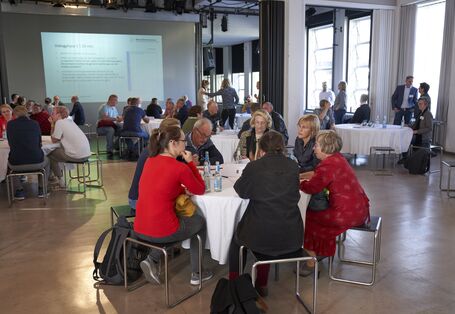 In einem großen Raum stehen mehrere runde Tischen, an denen diskutierende Menschen sitzen.