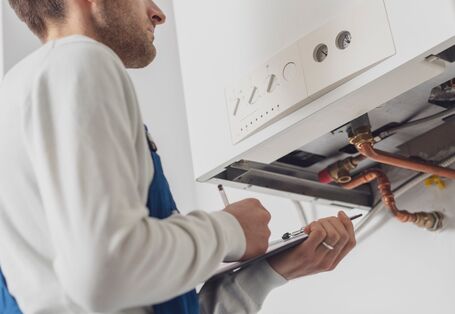 Ein Handwerker in blauer Arbeitskleidung und weißem Pullover steht vor einem geöffneten Boiler und notiert sich etwas auf einem Klemmbrett.