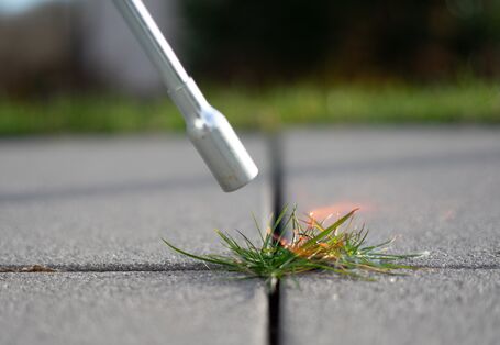 Ein Abflammgerät aus Metall brennt Gras in einer Fuge ab.