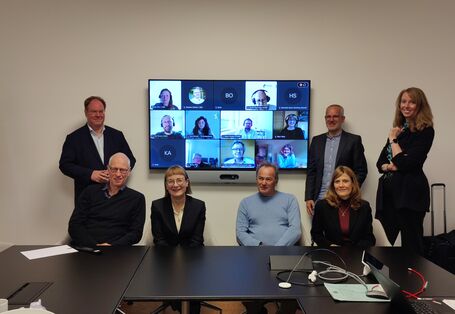 In a small meeting room, 7 people sit or stand in front of a screen. On the screen appear the faces of people in the meeting call.