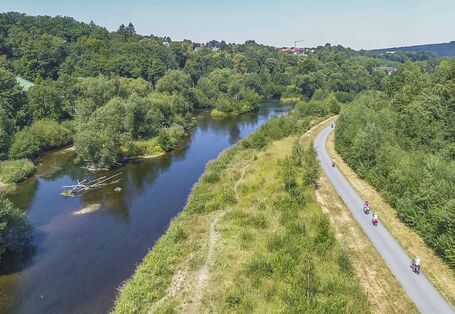 The river Ruhr near Arnsberg with numerous islands, gravel banks, dead wood and dense vegetation. Near the restored river runs a paved path on which several cyclists are on the move.