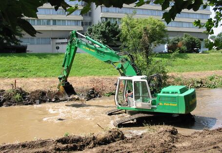 Foto: Ein Bagger bei der Bearbeitung der Ufer eines Fließgewässers. Am Ufer sind einzelne Bäume zu sehen. Im Hintergrund eine Brücke.