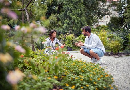 Zwei Personen hocken, ins Gespräch vertieft, in einer Gartenanlage.