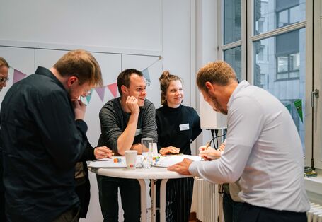Teilnehmende stehen an einem Stehtisch und arbeiten gemeinsam an vor ihnen liegenden Papieren. 