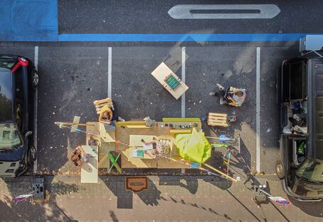 Auf einem Parkplatz steht ein großes Gefährt quer und Menschen sind aus der Vogelperspektive zu sehen, die im Freien an ihren Laptops arbeiten. 