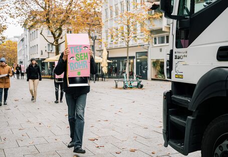Mann läuft duch Fußgängerzone und guckt durch das Tiefsehrohr, einem umgedrehten Periskop, und nimmt so die Perspektive von kleineren Menschen ein. 