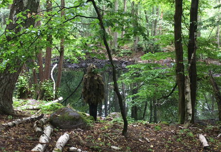 Fotografie eines Menschen, der mit Naturmaterialien verkleidet ist und im Wald vor einem Teich steht