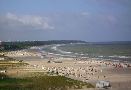 Blick auf Strand mit Strandkörben und touristischer Infrastruktur, im Wasser Kite- und Windsurfer. 