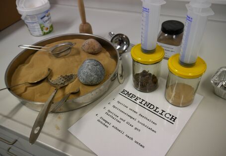 Auf einem Tisch stehen zwei Gläser mit Boden, in die mit jeweils einer großen Spritze Luft gepustet werden kann. Daneben steht feiner Sand, der durch ein Sieb rinnt.