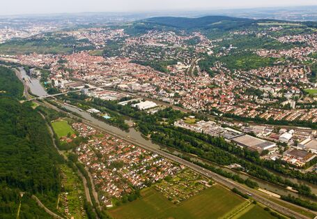 Luftbild einer Gemeinde in hügeliger Landschaft mit Fluss, Autobahn, Gewerbegebiet und Bahnstrecke im Vordergrund
